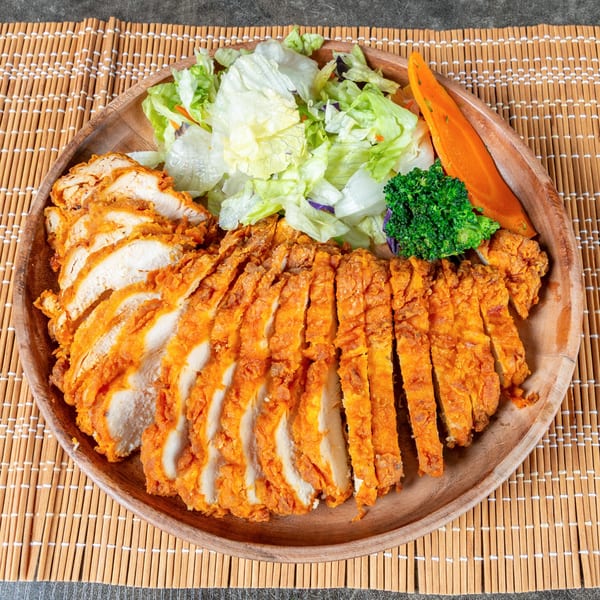 Sliced fried chicken with a side of fresh green salad, broccoli, and a carrot slice on a wooden plate.