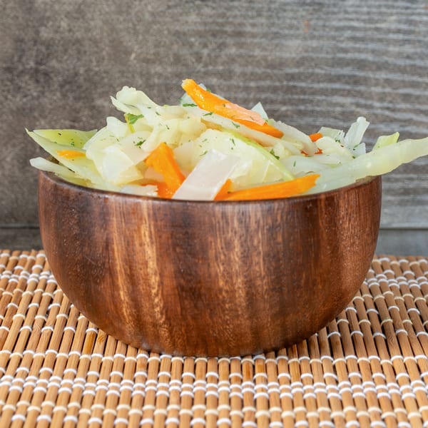 A wooden bowl filled with Jamaican steamed cabbage and carrots, garnished with herbs, on a bamboo mat.
